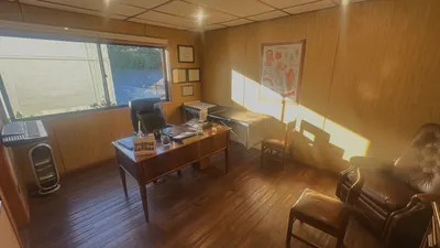 Consulta con paredes de madera: escritorio, camilla, lámina de la laringe, un sillón de cuero y sillas, con sol entrando por la ventana.